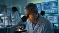 Senior male scientist examining sample through microscope in modern laboratory with digital displays and research team working in background.