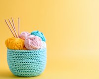 Colorful yarn balls in pink, yellow and turquoise placed in handmade crochet basket against vibrant yellow background, knitting needles included. Perfect for craft projects.
