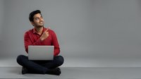 Young man with dark hair and South Asian ethnicity wearing a red button-up shirt, sitting cross-legged while using a laptop. Suitable for business, technology, remote work, and lifestyle concepts.