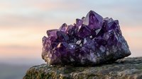 Large raw amethyst crystal cluster on a rocky surface with a soft pastel sunset sky in the background, showcasing natural purple gemstone beauty.