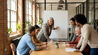 Diverse team of professionals collaborating at wooden table during business meeting in modern loft office with brick walls and large windows, suitable for corporate communications and workplace concepts.