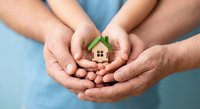 Family hands protecting small wooden house model symbolizing home insurance, real estate investment, mortgage protection and property security concept.