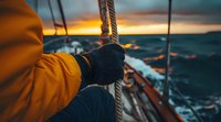 Gloved hands gripping rope on sailing vessel during dramatic ocean sunset, waves crashing against boat with orange sky and stormy waters in background.