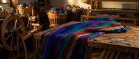 Colorful woven textile blanket draped over rustic wooden table in traditional craft workshop with spinning wheel and handmade baskets in warm ambient lighting.