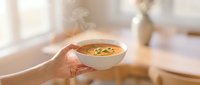 Hands holding warm bowl of creamy soup with herbs in cozy kitchen setting. Comfort food concept for healthy eating and home cooking lifestyle.