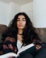 Young woman with long dark hair wearing ethnic pattern cardigan relaxing on couch while reading book. Natural lighting creates soft atmosphere for lifestyle portrait.