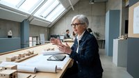 Senior female architect with gray hair and glasses reviewing blueprints at modern office desk under skylight, professional woman in business attire working on construction plans.