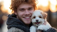 Smiling young man with curly reddish-brown hair holding small white fluffy dog outdoors during golden hour sunset, both looking at camera.