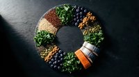 Colorful superfood ingredients arranged in circular pattern on dark slate background featuring quinoa, kale, blueberries, nuts and seeds for healthy nutrition.