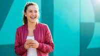 Young Caucasian businesswoman in pink blazer smiling while holding smartphone against modern turquoise office building background for corporate communication.