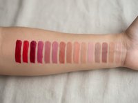 Lipstick swatches on woman's arm showing gradient from deep red to nude pink shades for cosmetic testing and color comparison purposes.