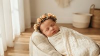 Peaceful newborn baby girl sleeping in cream wrap with floral headband in soft natural light studio setting for maternity photography sessions.