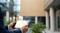 Young professional man in business attire holding tablet device outdoors near modern office building, suitable for corporate technology and business solutions marketing.