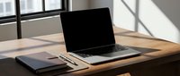 Laptop with blank screen on wooden desk beside notebook and pen, sunlight casting shadows, modern workspace setup for business and remote work.