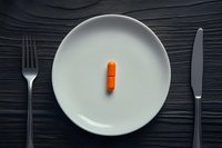 Orange pill capsule centered on white ceramic plate with dark silverware on black wooden table background, minimalist medical or dietary concept.