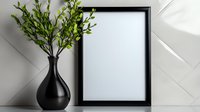 Black vase with fresh green leaves next to empty black photo frame on white wall background with natural sunlight and shadows, minimalist home decor setup.