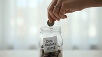Hand dropping coin into glass jar labeled our dream home filled with money for mortgage savings and real estate investment planning.