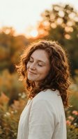 Young Caucasian woman with curly brown hair enjoying peaceful moment outdoors during golden hour sunset with eyes closed and gentle smile in natural setting.