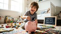 Young boy, approximately 5-8 years old, Caucasian ethnicity, inserting coin into pink piggy bank in bright bedroom surrounded by toys and books, with savings motivation sign displayed.