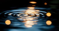 Rippling water surface with golden bokeh lights creating concentric circles, reflecting warm glowing orbs against dark background for tranquil spa atmosphere.