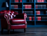 Luxurious burgundy leather armchair with brass studs against dark library shelves filled with red books creates atmospheric interior scene for law office or study.