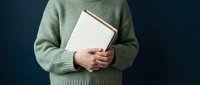 Person in cozy green sweater holding open book against dark background for reading and literature concepts.
