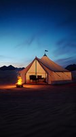 Luxurious glamping tent illuminated at dusk in desert landscape with campfire and mountain silhouettes. Cozy outdoor accommodation under starry twilight sky.