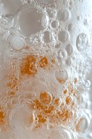 Macro shot of fizzy bubbles in carbonated orange drink against light background, showing delicate foam texture and effervescent spheres in soft lighting.