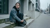 Young woman sitting alone on urban steps wearing denim jacket and dark clothing, expressing loneliness and contemplation in modern city environment.