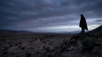 Silhouette of person standing on rocky outcrop overlooking vast desert landscape under dramatic stormy sky at dusk with purple and gray clouds.