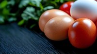 Fresh organic eggs in various natural shades from brown to white arranged on rustic wooden surface with green leafy vegetables in background for cooking.