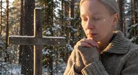 Young blonde woman praying peacefully beside wooden cross in snowy winter forest during golden hour sunlight for spiritual meditation and faith concepts.