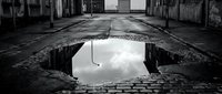 Abandoned urban street with large pothole filled with water reflecting cloudy sky, depicting decay and infrastructure damage in derelict industrial area.