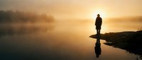 Silhouette of person standing on rocky shore at misty lake during golden sunrise with perfect water reflection creating peaceful solitude moment.
