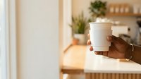 Hand holding white takeaway coffee cup in modern kitchen with wooden shelves and potted plants, suitable for cafe, beverage, and lifestyle marketing.