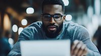 Young African American businessman in glasses working on laptop in evening office, focused on screen with bokeh lights in background. Professional remote work concept.