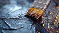Macro shot of paintbrush with brown bristles applying clear varnish or paint on dark textured surface with water drops, creating artistic reflective effect.