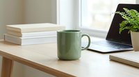 Green ceramic mug on wooden desk with stacked books, laptop, and potted plant near window for home office workspace.