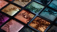 Crushed metallic eyeshadow palette featuring vibrant shimmering colors from pink to turquoise, brown and purple with detailed texture of broken powder makeup.