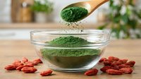 Green matcha powder falling from wooden spoon into glass bowl with goji berries on wooden surface, kitchen background.