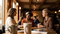 Three diverse young professionals having casual business meeting in modern coffee shop with warm lighting and wooden interior design.