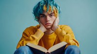 Young man with colorful blue and yellow hair wearing mustard sweater reading book while resting chin on hands against turquoise background for education concepts.