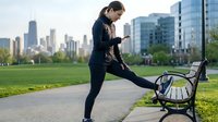 Young woman of East Asian descent in athletic wear stretching leg on park bench with urban skyline background, fitness and wellness concept for exercise routines and outdoor training.