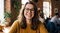 Happy young Caucasian woman with glasses and yellow sweater smiling in modern office or cafe workspace with brick walls and plants in background.