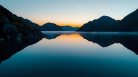 Tranquil mountain lake at dawn with perfect mirror reflection of silhouetted peaks against orange sunrise sky, creating symmetrical landscape composition.