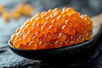 Fresh red caviar in black ceramic spoon on dark stone background, macro shot showing detailed texture of salmon roe pearls in natural lighting.