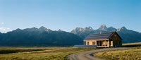 Isolated yellow farmhouse with curved driveway set against dramatic mountain range under clear blue sky, suitable for rural lifestyle, travel, and real estate design projects.