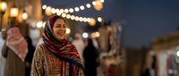 Young Middle Eastern woman in traditional headscarf smiling at evening market with warm bokeh lights creating festive atmosphere for cultural celebrations.