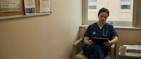 Asian female healthcare worker in blue scrubs with stethoscope reading tablet in medical office break room during shift.