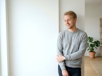 Young Caucasian male in casual gray sweatshirt leaning against white wall in modern interior space with potted plant, suitable for lifestyle and home decor content.